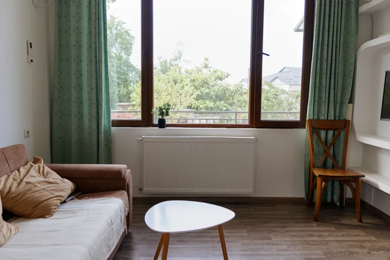 A bright, cozy living room with a white sofa, wooden coffee table, and green curtains under large windows. Natural light emphasizes soft textures and casual home lifestyle.