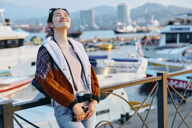 Woman in stylish fur jacket posing by harbor with boats and camera, enjoying sunny day and ocean breeze, travel fashion lifestyle by waterfront