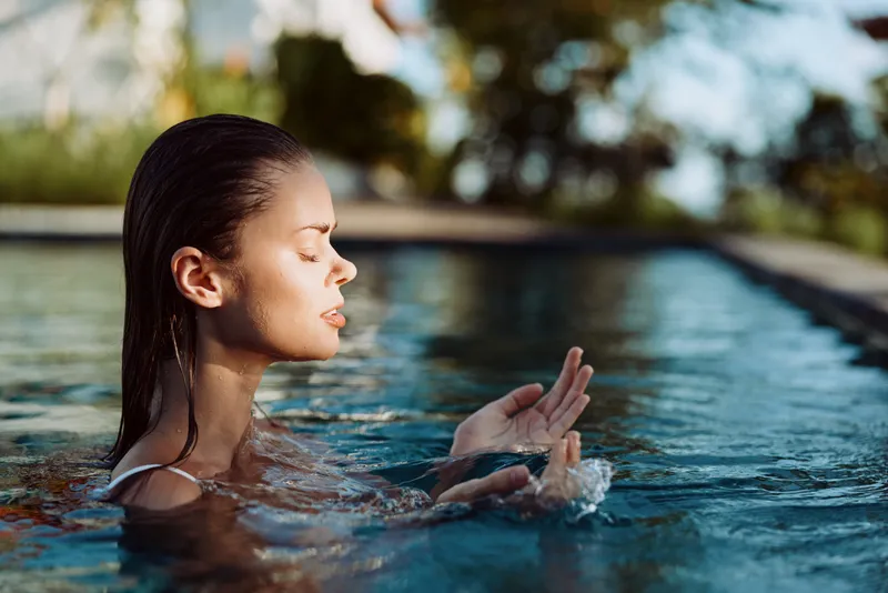 Young woman with wet hair relaxing in a swimming pool outdoors. She holds water in her hands with closed eyes, enjoying a peaceful moment in natural light. Lifestyle concept.