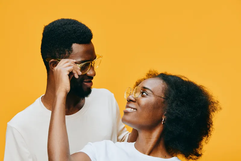 Young African couple smiling and wearing glasses against a bright yellow background, conveying love and happiness in a stylish, modern setting