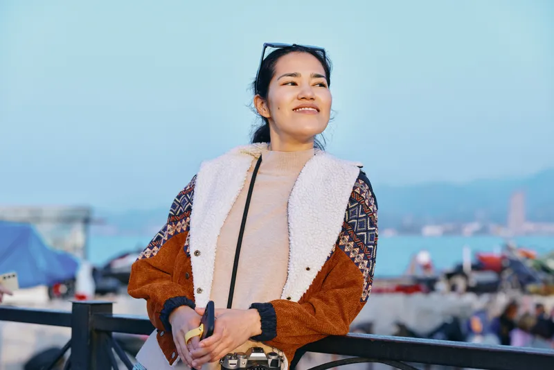 Smiling woman in a stylish jacket enjoying a breezy seaside day, sunglasses on head, casual fashion on a boardwalk with ocean backdrop and distant boats, candid lifestyle moment.