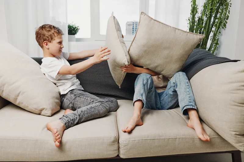 Two playful children having fun on a beige sofa with pillows in a bright living room. Concept of childhood joy, sibling interaction, and indoor playtime at home.
