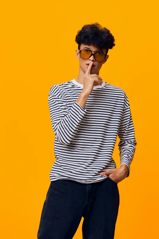 young man wearing striped shirt and sunglasses with curly hair making silence gesture against bright orange background expressing secrecy and calm mood