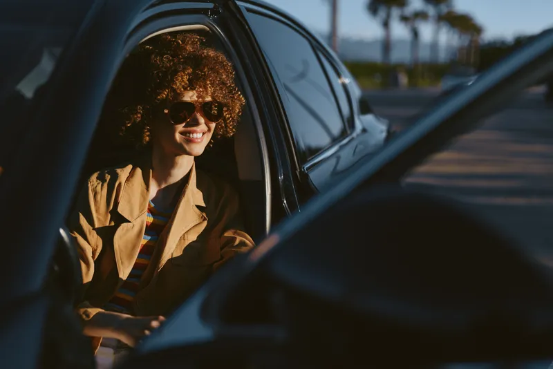 Smiling woman with curly hair sits in a car, sunlight on the interior, retro film color palette, lifestyle moment, warm and cheerful vibe, candid travel portrait.