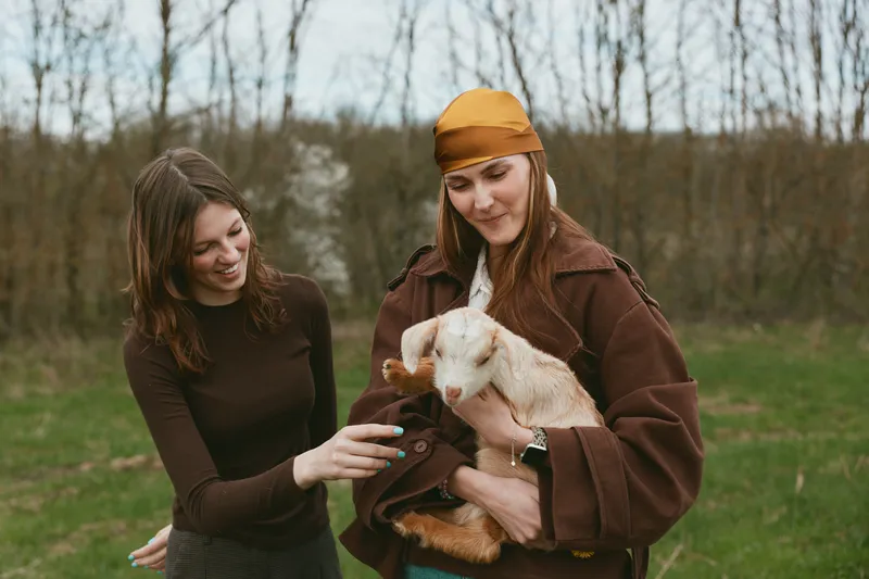 Two women outdoors with a goat kid, sharing a warm moment. Casual outfits, natural light, rustic setting. A lifestyle scene focused on friendship, nature, and gentle interaction with animals.