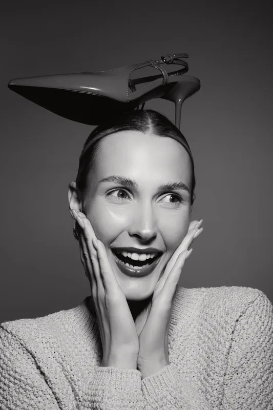 Happy Caucasian woman with light skin smiling, holding face with hands and a high heel shoe on head, isolated on grey background.