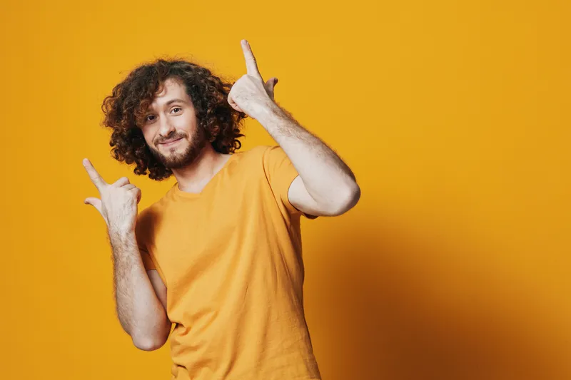 Young man with curly hair in a cheerful yellow shirt poses against a bright yellow background, expressing joy and positivity with hand gestures