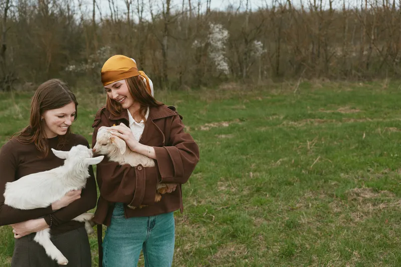 Two young women enjoy a spring day outdoors while holding and petting a white baby goat in a grassy field with bare trees in the background