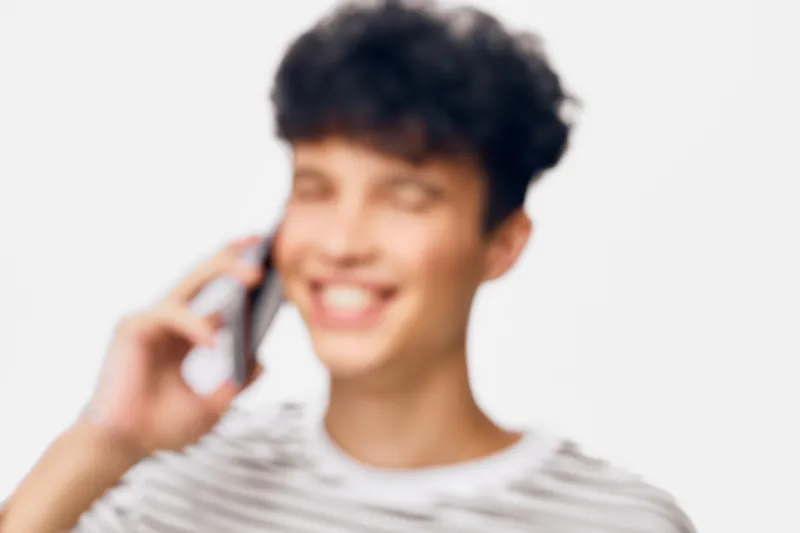 smiling man talking on phone, young adult male with curly hair wearing striped shirt, bright background with soft focus, cheerful communication concept