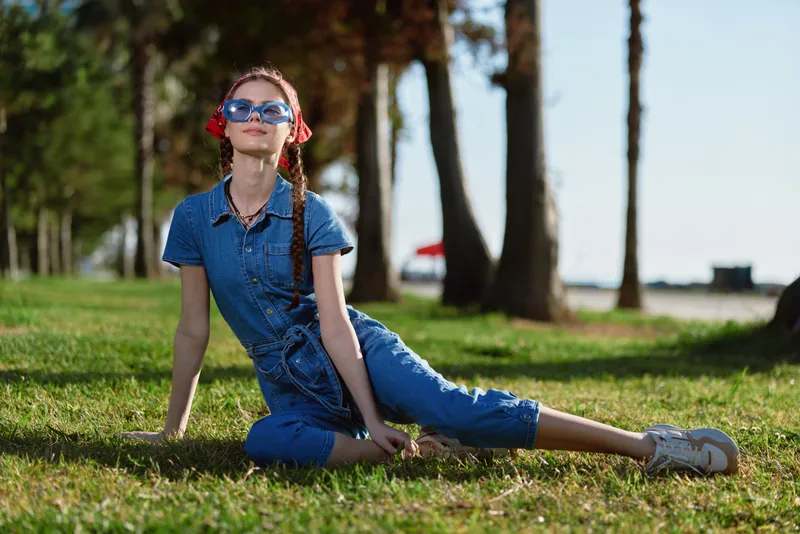 Young woman in blue jumpsuit relaxing on grass with sunglasses under bright sunny sky near palm trees, enjoying warm weather and casual outdoor style.