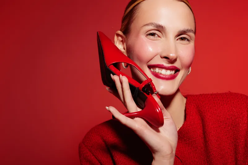Smiling woman in red sweater holds red high heel shoe near face on red background. Concept of fashion, style, beauty, and vibrant color contrast in studio portrait.