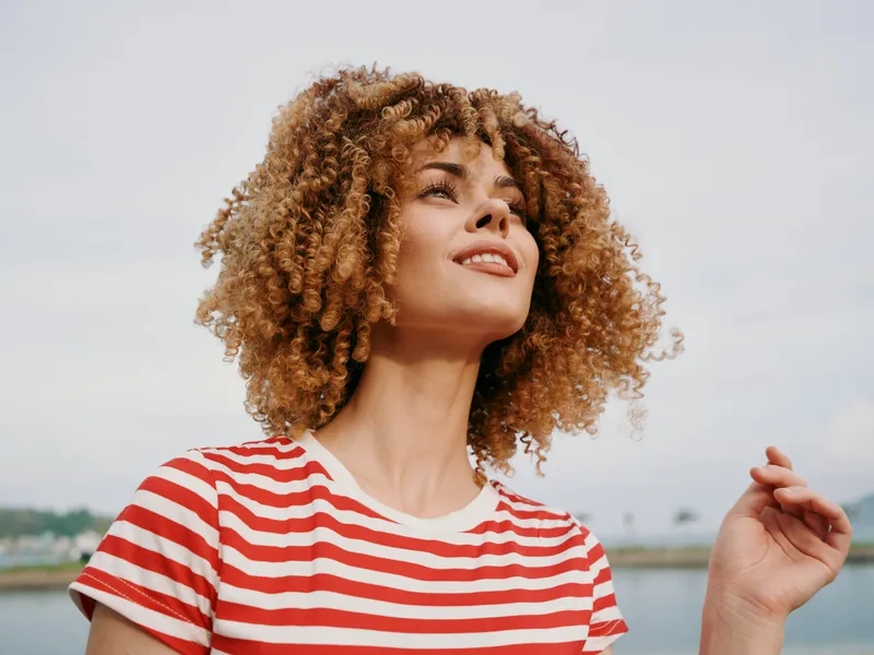 Portrait of a young woman with voluminous curly hair wearing a red and white striped tee, smiling softly as a light breeze lifts her curls against a bright sky.