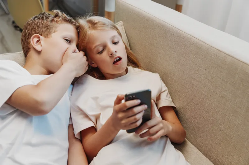 Two children sitting on sofa whisper secrets while holding smartphone, sharing surprise and curiosity in casual home environment.