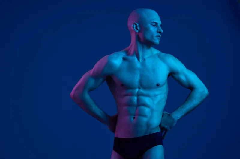 A muscular man posing in dramatic lighting, showcasing his physique. The image features a striking blue color palette that enhances his defined muscles.