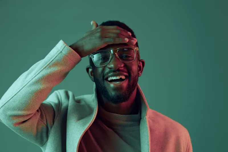 Smiling young Black man in glasses touching his forehead, wearing a stylish coat against a vibrant green and pink background, exuding confidence and joy
