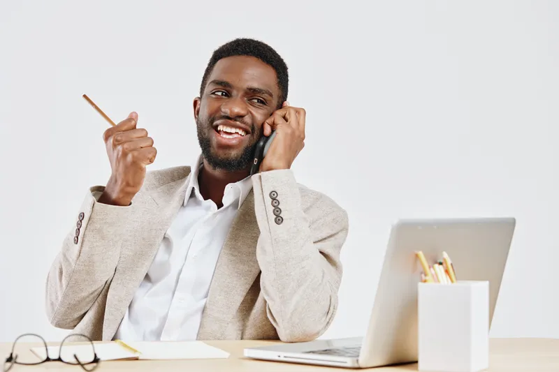 Smiling man with dark skin, cheerful expression, talking on phone, sitting at desk with laptop, holding pen, casual beige blazer, bright modern office background, lifestyle concept.
