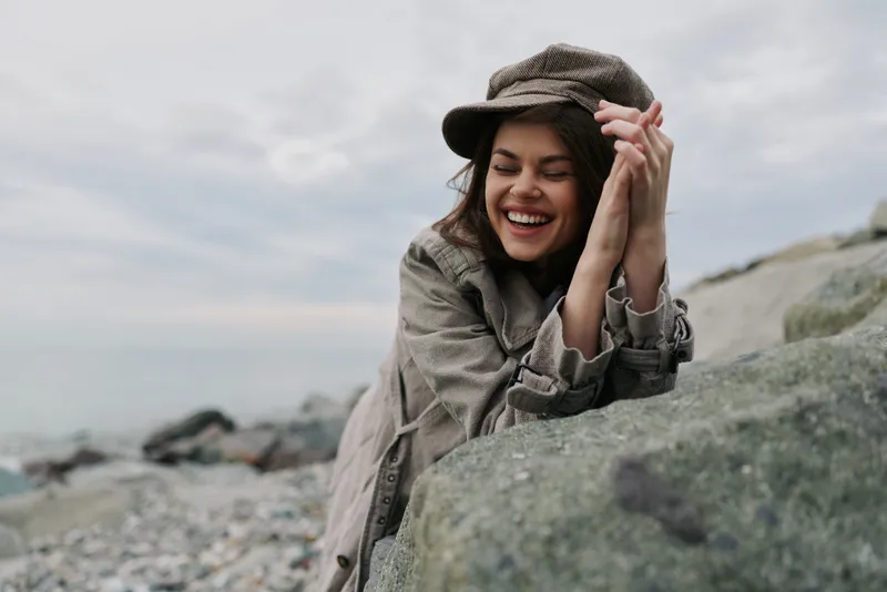 Smiling woman wearing a checkered coat and matching hat leans on a rock by the sea shore. Overcast sky and rocky beach create a calm, natural outdoor atmosphere of joy and relaxation.