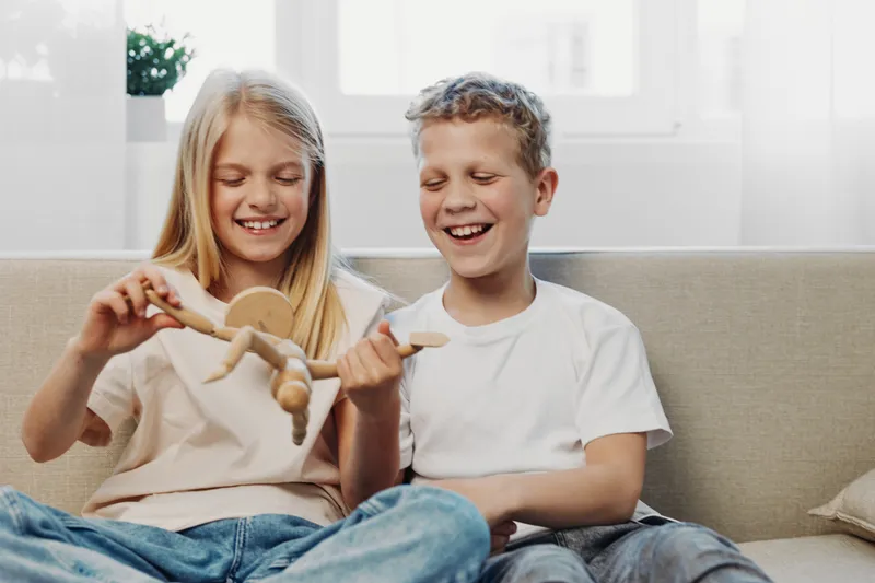 Two happy children play with a wooden doll on a couch in a bright living room. The boy and girl smile and enjoy creative imaginative play together at home.