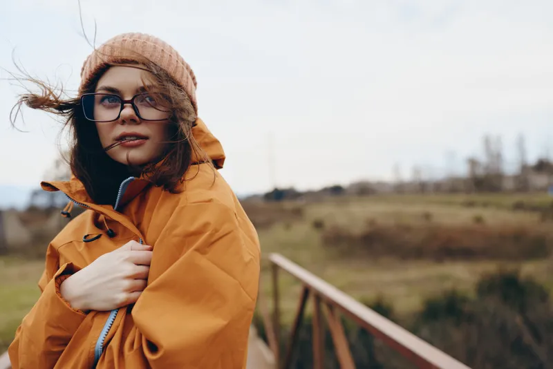 Woman lifestyle outdoors in winter wearing orange coat and knit hat portrait with wind blowing hair soft sunlight smile scenic field countryside background natural colors