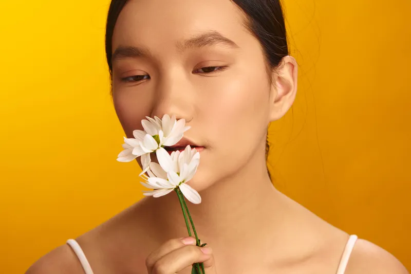 asian woman softly smells a white flower in a bright studio, close up portrait with warm yellow backdrop, serene mood and gentle elegance