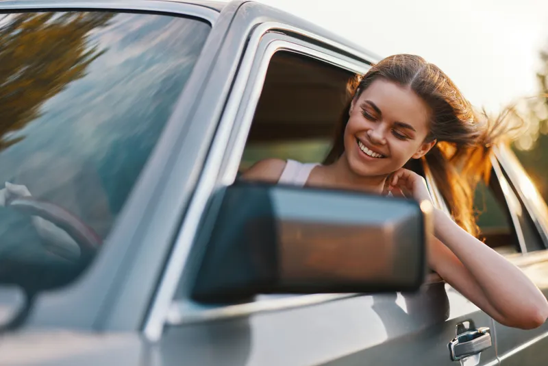 Happy young woman enjoying a road trip in a vintage car, with wind in her hair, bright sunset background. Perfect for travel and adventure themes.
