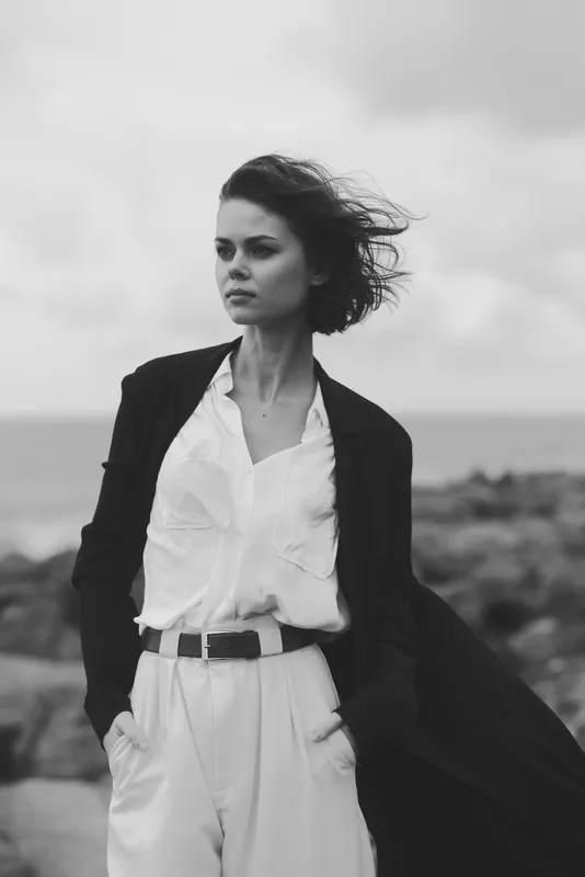 Stylish woman stands in a windy coastal setting wearing a tailored blazer and white shirt, captured in black and white with a dramatic sea breeze and confident stance in frame.
