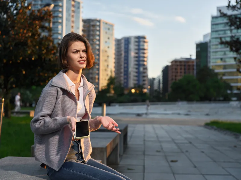 A cheerful woman stands outdoors in an urban park, texting on a smartphone in a modern city setting. She smiles, projects confidence, and radiates a dynamic business vibe.