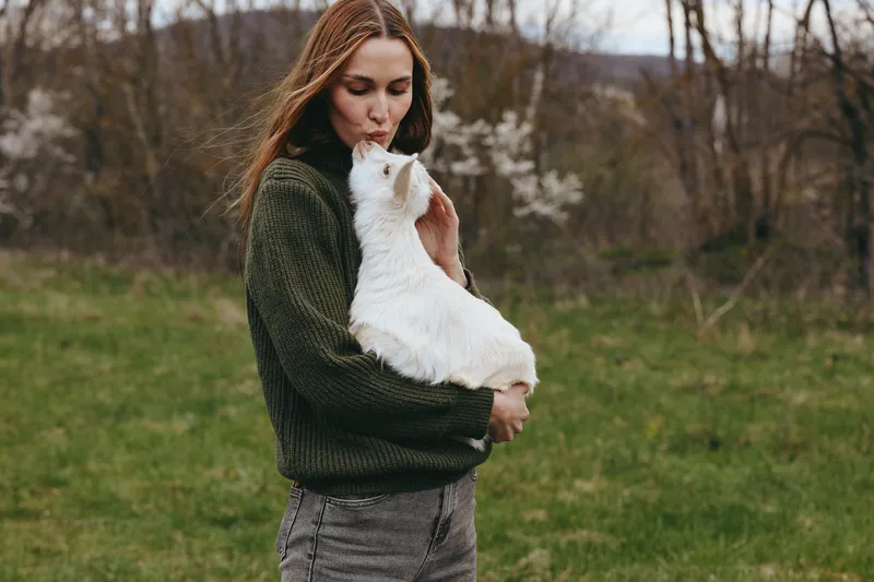 Young woman with long brown hair kisses a white baby goat while standing outdoors in a green knitted sweater and patterned pants during a calm spring day