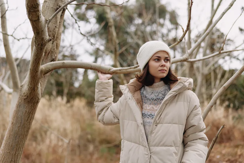 Woman wearing a beige puffer coat and knit beanie stands among leafless trees in a frosty landscape, gazing into the distance with calm, seasonal mood.