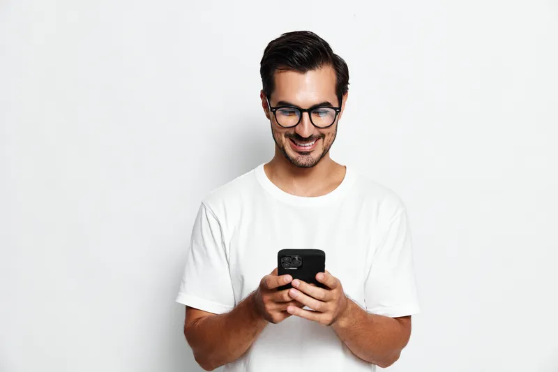Smiling happy cheerful young man wearing white t-shirt looking at mobile cell phone, isolated on plain solid light background. People lifestyle concept.