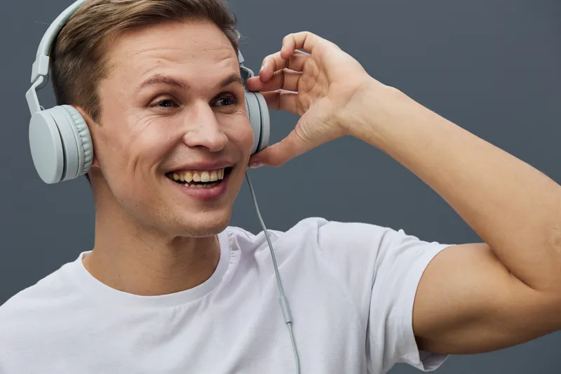 Happy young caucasian man wearing large over-ear headphones listening to music or audio, smiling excitedly, isolated on plain gray background, casual white t-shirt, studio portrait. People lifestyle c