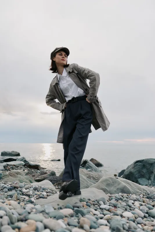 Young woman in trench coat and cap stands confidently on rocky beach with cloudy sky and calm sea in background. Concept of fashion, confidence, and outdoor lifestyle.