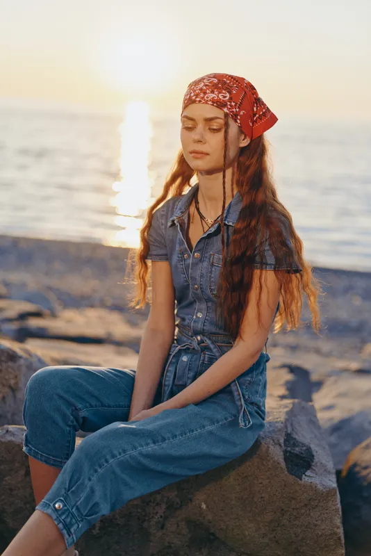 Young woman meditating by the sea during sunset with long hair, casual denim outfit, and red bandana, peaceful and serene coastal scene with warm light.