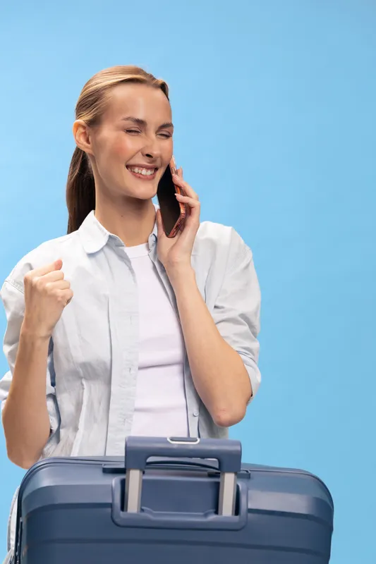 Happy woman with suitcase clenches fist and talks on phone against blue background. Concept for travel, success, communication, and positive emotions in lifestyle and vacation.