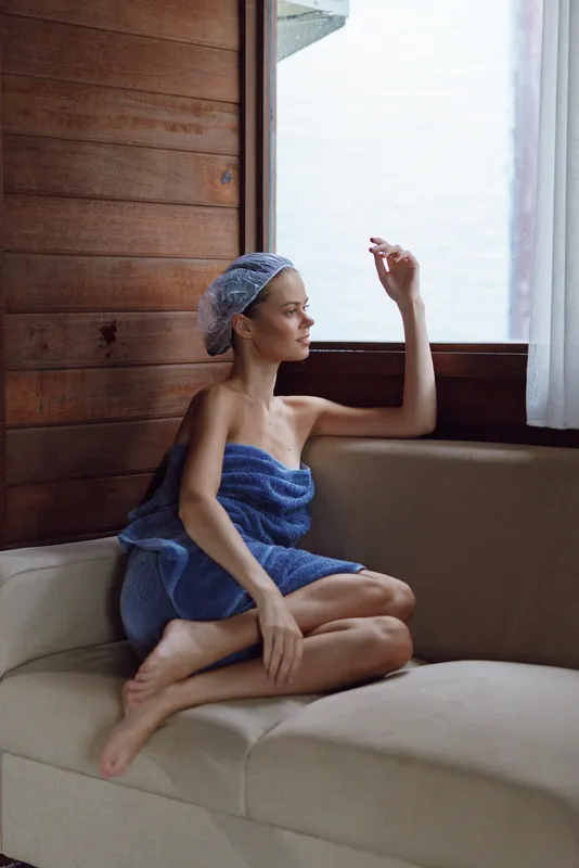 woman relaxing indoors wrapped in a blue towel with shower cap, sitting on beige sofa near wooden wall and window, natural light creating calm atmosphere