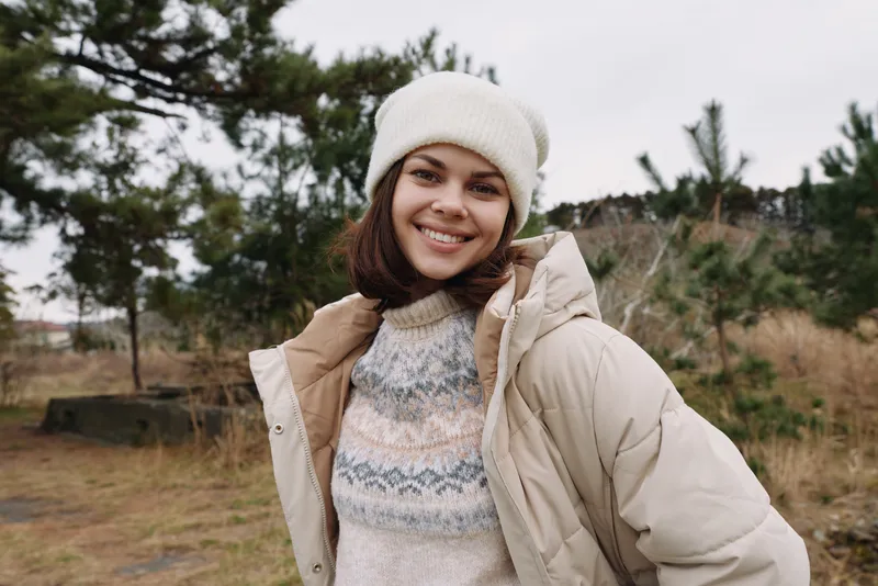 Smiling woman wearing a beige puffer coat and patterned knit sweater outdoors in a chilly landscape with trees and open space.