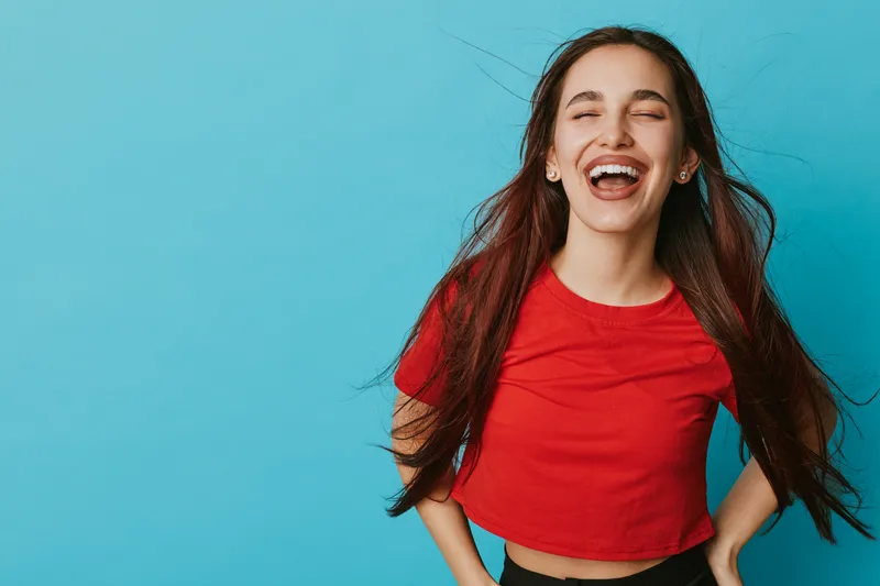 Young woman with long hair laughing and standing against blue background wearing red shirt. Happy female portrait with closed eyes and natural smile in casual style and expression.
