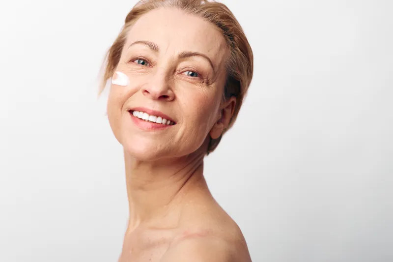 Smiling mature woman with natural skin applies white face cream on cheek, isolated on white background. Concept for skincare, aging beauty, self-care, wellness, and healthy lifestyle.
