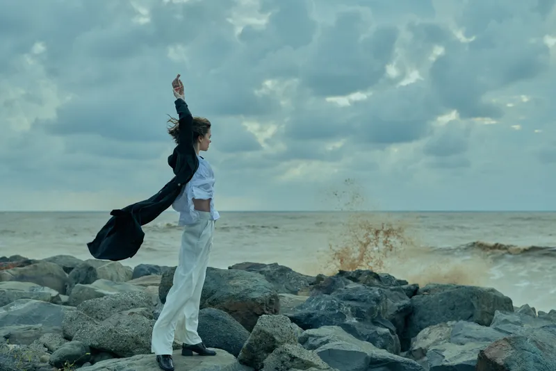 Seaside scene with a woman standing on rocks, flowing jacket and white outfit, wind sweeping the horizon, capturing dynamic movement against a cloudy sky