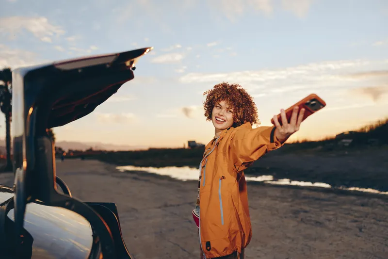 Smiling woman lifestyle near open car trunk at sunset, outdoors by a quiet road, curly hair, casual jacket, arms raised, travel moment in golden hour