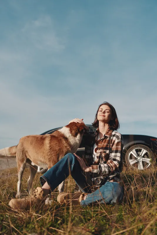Woman sits on tall grass outdoors with a dog in a bright field, casual plaid shirt and jeans, car in the distance, capturing a peaceful moment of companionship and nature.