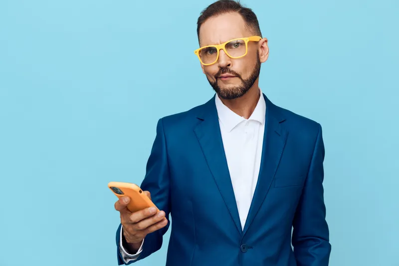 A professional man in a blue suit stands against an isolated blue background, exuding confidence, focus, and determination as he holds a smartphone and gazes ahead.