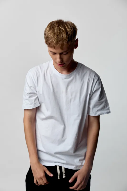 Young man, white t shirt, casual, contemplation, studio, fashion, relaxed posture and downturned gaze in a minimalist setting with neutral tones, soft shadows, simple composition conveying calm focus