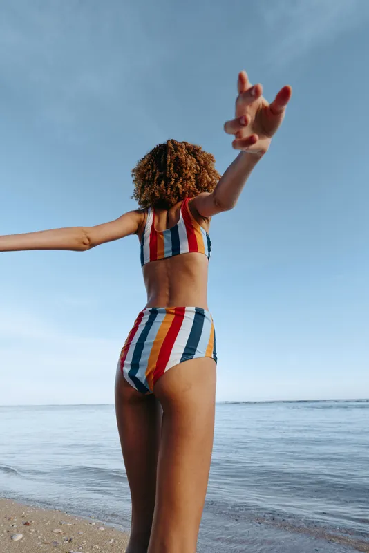 Curly haired woman wearing colorful striped swimsuit stands on sandy beach by the ocean under clear blue sky with arms lifted in playful pose, summer vacation mood, natural light