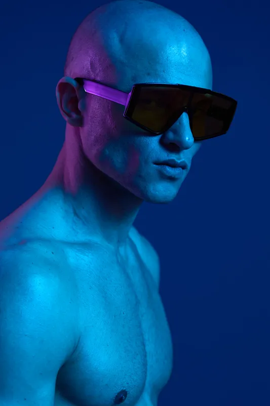 Stylish man with sunglasses poses against a vibrant blue background. The image showcases a striking look with a modern aesthetic, emphasizing contemporary fashion trends.