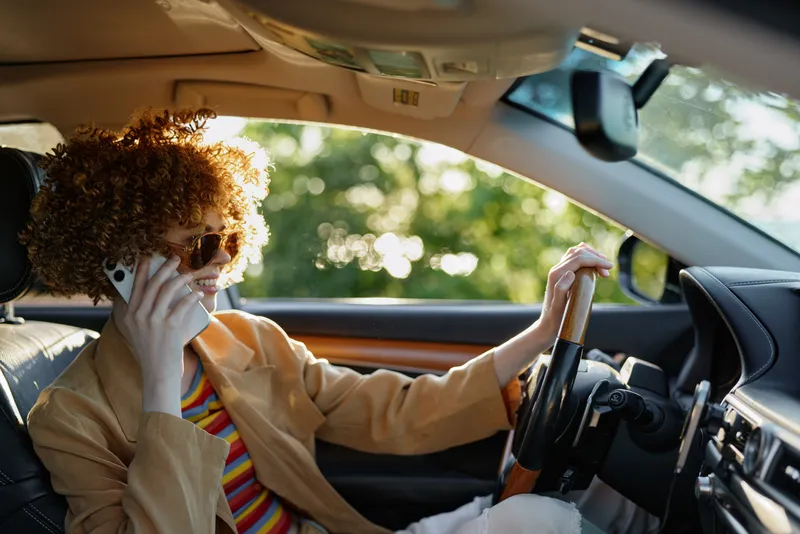 A stylish driver in a retro outfit talks on a phone inside a sunlit car, showcasing a vibrant lifestyle, bold color, and street fashion during a relaxed road moment.