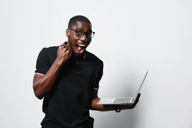 African American man with dark skin and glasses holding laptop celebrating success with excited expression on white background. Casual black shirt and positive emotion.