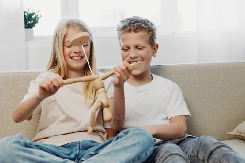 Two happy children play with wooden puppets on a couch in bright living room, enjoying creative and imaginative playtime together.