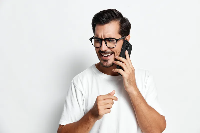 Happy excited young man wearing glasses talking on smartphone while smiling and gesturing with hand, isolated on plain white background. Lifestyle concept.