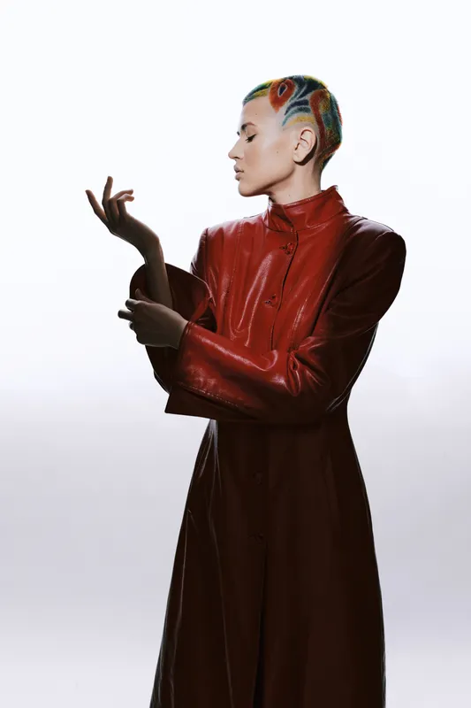 Woman with colorful shaved hair wearing a red leather coat poses elegantly against a minimalist white background with soft lighting.